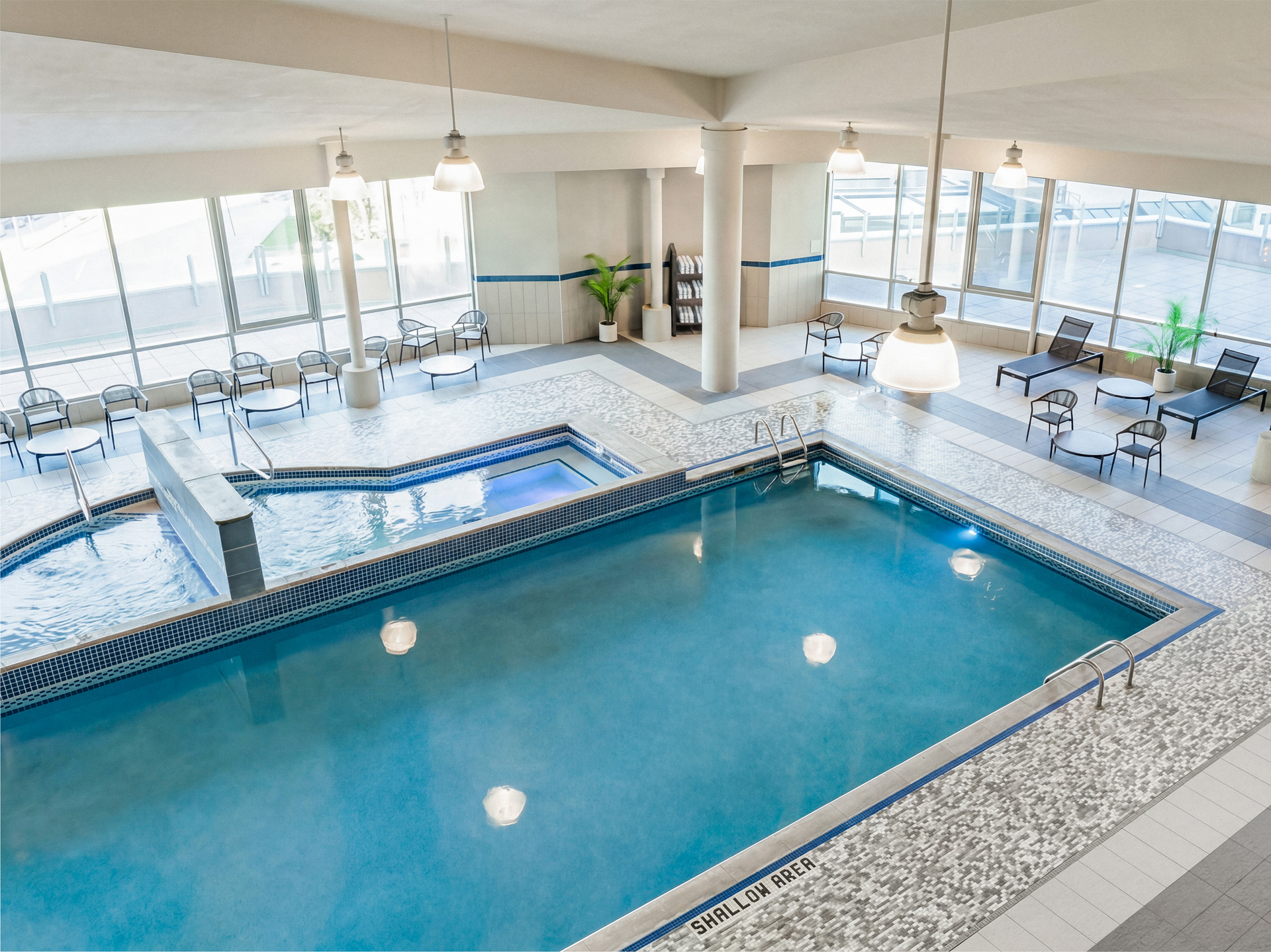 A large indoor swimming pool, with two hot tubs connected to the pool, large windows surrounding the pool deck with tables and lounge chairs.